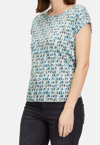 Košile s krátkým rukávem vyrobená z světle béžové látky s geometrickým vzorem v modré, hnědé a černé barvě. Měkká, hladká textury, kulatý výstřih.