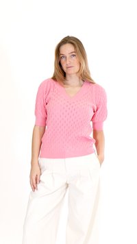 Žena s dlouhými blond vlasy, oblečená v růžovém texturovaném svetru s krátkými rukávy a bílých kalhotách s širokými nohavicemi, rukou v kapse, neutrální pozadí.