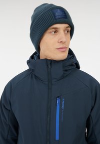 Marine waterdichte jas met hoge kraag, voorzien van een blauwe ritszak en contrasterend logo, gecombineerd met een gestructureerde, donkerblauwe gebreide muts.
