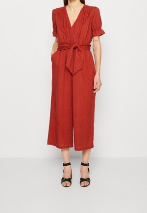 Kvinde iført en bredbenet jumpsuit i rust-rød med puffede ærmer, dyb V-udskæring, et bundet bælte i taljen og sorte sandaler med ankelrem.