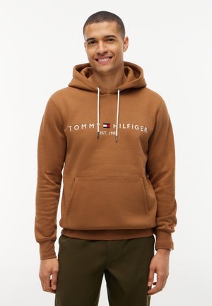 Homme souriant portant un sweat à capuche Tommy Hilfiger marron avec des cordons blancs et un pantalon vert foncé sur un fond blanc uni.