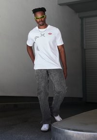 White t-shirt with black graphic design and red logo, paired with gray jeans. Model wearing green sunglasses and white sneakers.