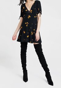 Svart klänning med V-ringning, korta ärmar och blommönster, kombinerad med knähöga svarta stövlar. Klänningen har en utsvängd kjol och knyts vid ärmarna.