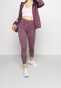 Sportovní outfit složený z světle růžového crop topu a tmavě fialových legín s tkaničkou, doplněný o fialovou mikinu na zip a bílé tenisky.