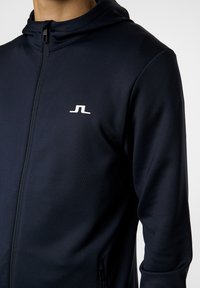 Felpa con zip di colore navy in tessuto morbido, con colletto alto, tasche laterali e un piccolo logo bianco sul petto.