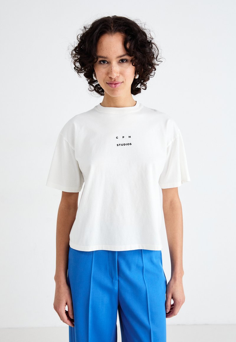 Hvid bomulds T-shirt med korte ærmer, prydet med en centreret sort tekstprint "C P H STUDIOS." Sammen med blå skræddersyede bukser.