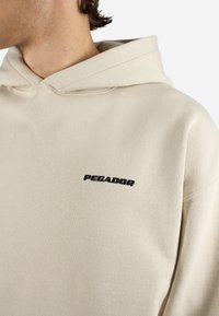 Beige hettegenser med snor i hetta og svart "PEGADOR" logo brodert på venstre bryst. Laget av mykt, teksturert stoff.