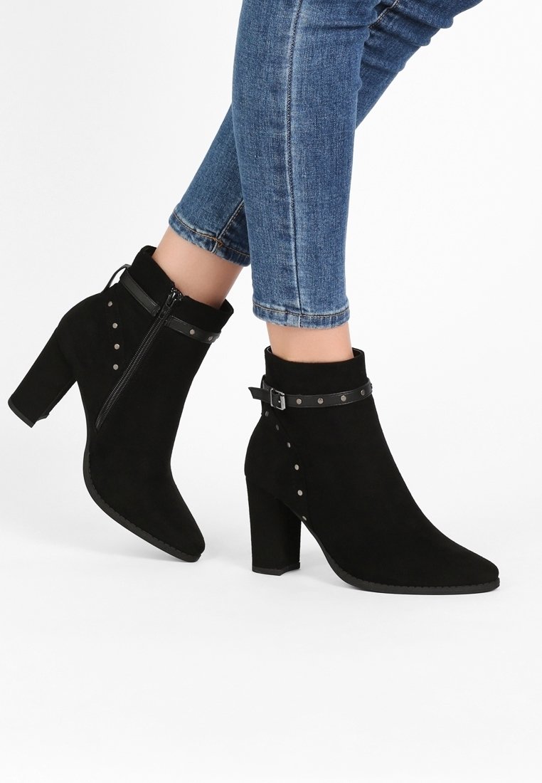 Bottines en daim noir avec talons carrés, fermeture éclair latérale, lanière à boucle et clous métalliques portées avec un jean skinny bleu.