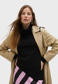 Beige trench coat with a textured finish over a black turtleneck sweater. The outfit features a pink and black striped skirt.