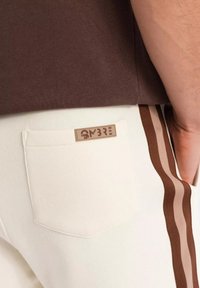 Un pantalon blanc avec une poche arrière ornée d'une étiquette marron. Des rayures marron et beige descendent sur les côtés, complémentant le haut marron foncé au-dessus.