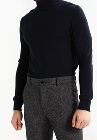 Man som bär en svart polotröja och högmidjade grå tweedbyxor med bälteshällor, stående mot en vit bakgrund.