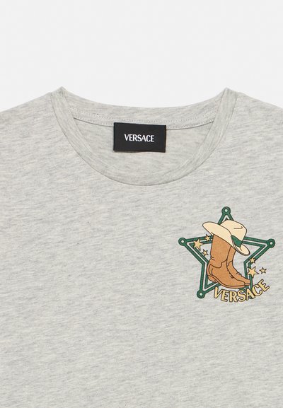 Ανοιχτό γκρι μπλουζάκι με στρογγυλή λαιμόκοψη, ετικέτα Versace και γραφικό με πράσινο αστέρι σερίφη, καουμπόικα μπότες, καπέλο και το κείμενο "VERSACE" στο στήθος.