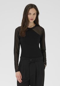 Mujer con cabello largo castaño, vistiendo un top negro con mangas largas de malla transparente y pantalones negros de cintura alta con un cinturón.