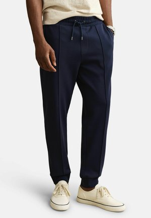 Homme portant un pantalon de jogging bleu marine avec des coutures frontales, une ceinture élastique avec cordon de serrage, des baskets beiges et une chemise à manches courtes beige clair.