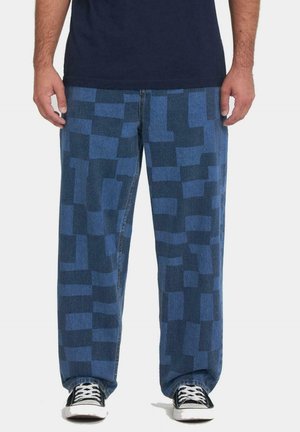 Hombre con pantalones azules sueltos con un patrón de bloques y zapatillas negras con cordones blancos sobre un fondo liso.