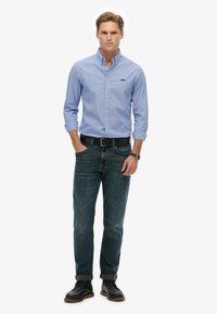 Lyseblå skjorte med lange ærmer, mørke jeans, sort bælte og sorte dress sko. Skjorten har et lille logo på brystet.