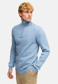 Jasnoniebieski sweter z prążkowanej dzianiny z kołnierzem na pół zamka, dopasowanymi rękawami i teksturowanym wykończeniem. Noszony z spodniami w neutralnym kolorze.