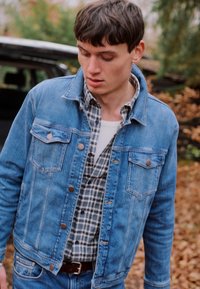 Jachetă din denim de un albastru deschis, cu două buzunare pe piept, închidere cu nasturi și o croială lejeră, purtată peste o cămașă din flanel cu carouri.