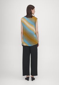 Ermeløs topp i blå, gul og beige bølgemønster; myk stoff med en flytende design. Kombinert med løse, svarte, lange bukser.