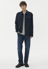 Mlad moški nosi mornarsko modro srajco s gumbi preko črtaste srajce, modre kavbojke in črne čipkaste čevlje, stoji z eno roko v žepu.