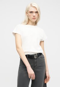 Jeune personne aux longs cheveux blonds portant une chemise blanche à manches courtes, un jean gris et une ceinture noire avec une boucle en forme de cœur.