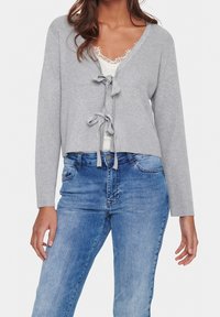 Grå rippad kort cardigan med långa ärmar och V-ringning, med en framknutning. Bärs över en vit spetsblus och blå denimjeans.