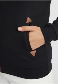 Juodas džemperis su priekiu kišene, puoštas trikampiais rudos odos akcentais. Audinys atrodo minkštas, o apačia turi briaunuotą tekstūrą.