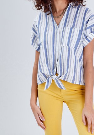 Femme portant une chemise à rayures verticales bleu et blanc nouée à la taille avec un pantalon moutarde et un collier chaîne en or.