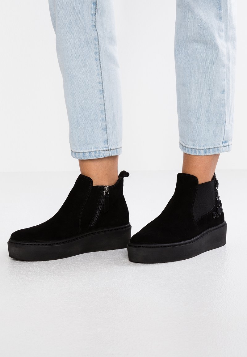 Černé semišové kotníkové boty s platformou, bočním zipem, elastickými vsadkami a dekorativními květinovými akcenty na boku.