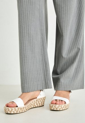 Personne portant un pantalon à rayures grises et des sandales à talons compensés blancs avec des semelles en jute, se tenant sur un sol clair.