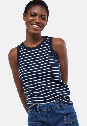 Personne souriante portant un haut sans manches rayé bleu marine et blanc ainsi qu'un pantalon en denim bleu, debout avec les mains derrière le dos.