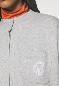 Närbild av en person som bär en grå knäppt jacka med en bröstficka där en vit Nike-logotyp syns och en orange polotröja under.