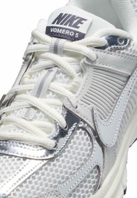 Baskets Nike Vomero 5 avec une tige en maille blanche, accents argentés, superpositions texturées et lacets plats blancs.