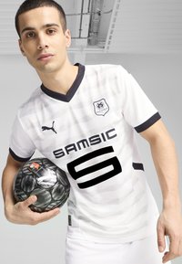Biely športový dres s čiernymi akcentmi, ktorý má výstrih do V, horizontálne pruhy a logo. Hráč drží futbalovú loptu s čiernym vzorom.