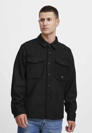 Black button-up shirt with two front pockets, made of textured fabric, featuring a collared neckline and long sleeves.