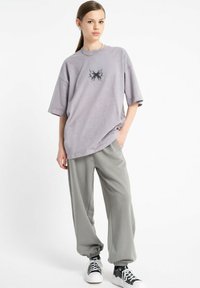 Młoda kobieta stoi, ubrana w oversize'owy fioletowy t-shirt z czarną grafiką, luźne szare dresy i czarno-białe trampki, na białym tle.