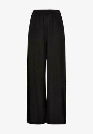 Vrouw die zwarte, wijde broek met hoge taille, teal crop top en zwarte platformsandalen draagt, staand in zijprofiel op een grijze achtergrond.