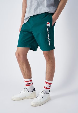 Pantaloni da nuoto turchesi con vita elastica, logo "Champion" bianco, abbinati a sneakers bianche e calze a righe rosse. Tessuto dalla texture liscia.