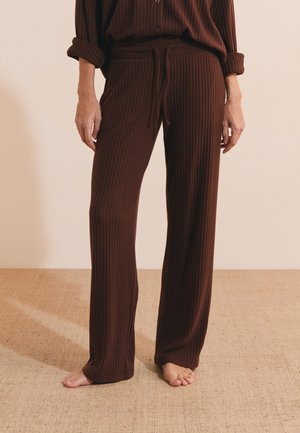 Donna che indossa pantaloni ampi in maglia a coste marroni e top abbinato, in piedi scalza su un pavimento beige testurizzato.