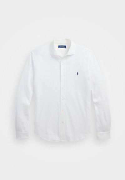 Λευκό πουκάμισο με μακριά μανίκια και κουμπιά, που διαθέτει κυρτό γιακά, υφή και ένα μικρό ναυτικό λογότυπο στο αριστερό στήθος. Κίτρινα κουμπιά.