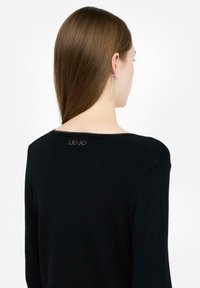 Mujer con cabello castaño liso que lleva una camiseta de manga larga negra con un pequeño logotipo "LIU·JO" en la parte posterior del cuello y pendientes de strass colgantes.