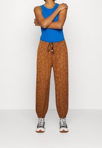 Niebieski top bez rękawów w połączeniu z luźnymi, brązowymi spodniami w wzory, które mają elastyczny pas z troczkiem i elastyczne mankiety, uzupełnione czarno-białymi trampkami.