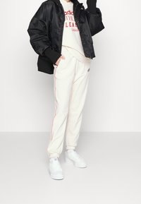 Zwarte satijnen bomberjack met geribbelde manchetten, witte rondehals sweatshirt met rood logo, crème joggers met rode bies en witte sneakers.