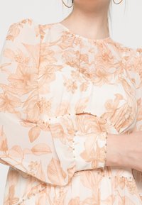 Blus med blommönster i ljus kräm och persika, med puffärmar och guldpärlor längs manschetterna och midjesömmen.