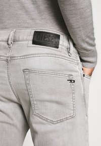 Vaqueros de mezclilla gris con un parche de cuero negro texturizado etiquetado 'DIESEL'. Las características incluyen bolsillos traseros cosidos y un acento sutil cerca de la cinturilla.