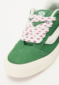 Grønne semskede sneakers med hvite detaljer, med rosa og hvite mønstrede snører og en rund, teksturert gummisåle.