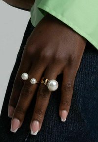 Guldring med tre vita pärlor i olika storlekar; fäst vid en hand med ljusa naglar med fransk manikyr. Grön ärm i bakgrunden.