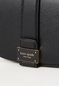Detailní záběr na popruh černé kožené kabelky se zlatým štítkem s nápisem "kate spade new york" na texturovaném koženém povrchu.