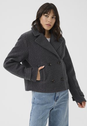 Femme portant une veste en laine gris foncé texturée, croisée avec de larges revers, et un jean bleu clair, debout devant un fond uni.