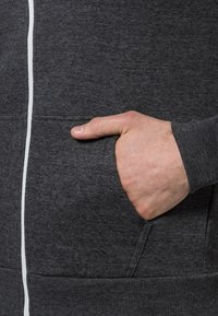 En vuxenhand är inskjuten i framfickan på en mörkgrå zip-up hoodie med vit dragkedjedetalj.